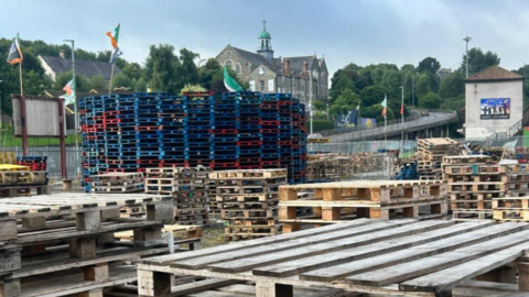 Shows large piles of pallets being stacked in preparation for a bonfire. Flags, including the Irish tricolour and Palestinian flag, are seen in the background.