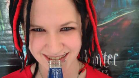 A close-up portrait shows Sophie Lancaster with black-and-red dreadlocks, facial piercings and a red top, holding the neck of a glass bottle between her teeth. A dark, fantasy-themed Harry Potter poster is visible in the background.