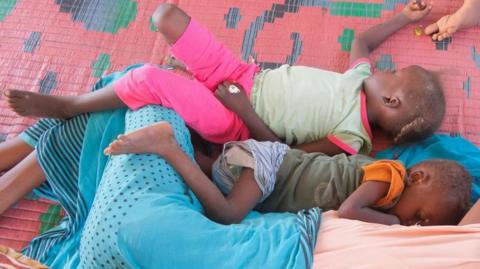 Two small children lie side-by-side on a pink grass mat next to someone covered in a cloth.