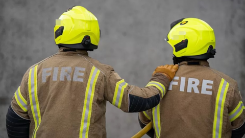 Two firefighters in mainly brown uniform, seen from the back. They are wearing yellow helmets and the firefighter on the left has a hand on a shoulder of the firefighter on the right.