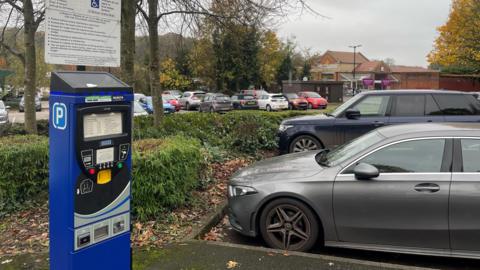 A pay and display machine in a car park. Cars are parked on the right and in the distance on the other side of some trees and bushes.