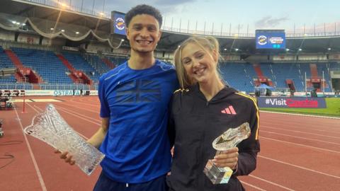 Joel Clarke-Khan, with short dark hair and wearing a blue T-shirt, with a black Union Flag on it, holds a glass trophy in his right hand while his left is behind Molly Caudrey - Molly has a dark blue tracksuit with yellow piping and holds a trophy shaped like a boot, made of glass, in her left hand. They stand on an athletics track.
