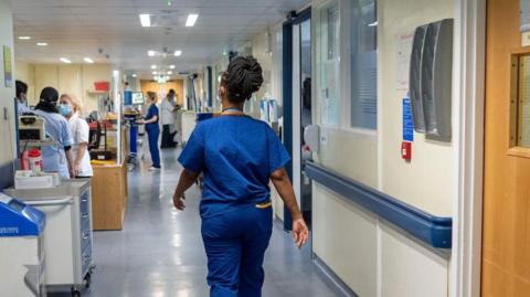 A nurse wearing blue scrubs walks down a hospital corridor. 