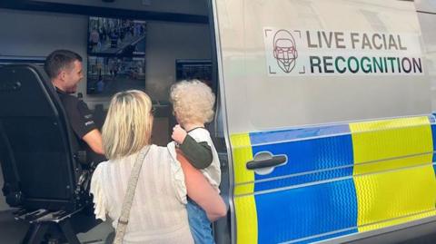 A policeman in a live facial recognition van, with a woman and child looking at screens, with their backs away from the camera. There are a number of screens in the van, and it has writing on the side. The policeman is smiling. 