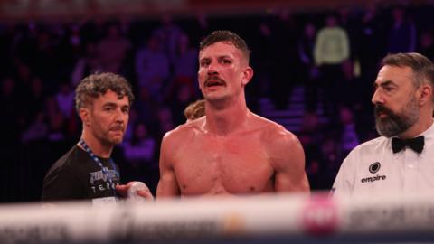 Nathan Heaney is stood topless in a boxing ring, with officials either side of him and a large crowd in the background behind.