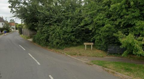 A sign for Chilton Road stands on the grass, surrounded by trees and shrubs. A winding road, with some old white houses can be seen in the distance.