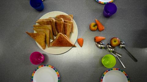 A grey table that has a plate of toast in the middle as well as brightly-coloured plastic cups and white plates with a colourful edge. There are some apples and carrots on the table too.