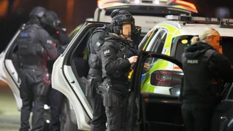 Four armed police officers standing behind police vehicles. There is also a woman with a police vest on in the foreground.