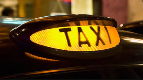 A large, yellow taxi light on top of a black cab is seen close-up in the image.