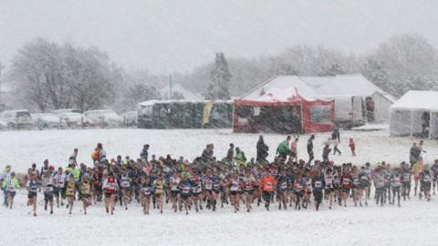 A crowd of people running in the distance on a field covered in snow. Snow is coming down heavily around them. The cars parked in the distance are completely covered. There are tents and portaloos at the back of the field.