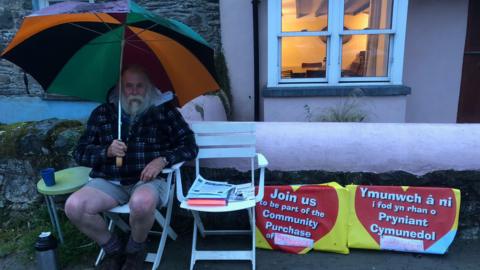 John Harries sitting under an umbrella next to posters saying "Join us to be part of the Community Purchase of Bethlehem"