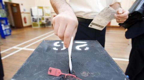 A hand places a ballot paper into a black ballot box at a polling station. The box has a numbered label on its lid and a red security seal attached to the slot, with a voter holding paperwork standing behind it in a hall.