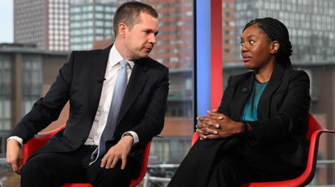 Robert Jenrick sits in a red chair on the left and Kemi Badenoch in a chair on the right. Jenrick turns to Badenock to speak to her.