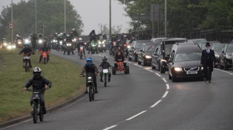 Dozens of bikers and youngsters on scrambler bikes ride alongside the funeral cortege. The bikes and cars have their lights on in the gloom. 