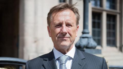 Martin stands infront of the county council building in Stafford on a sunny day, smiling at the camera. He is wearing a black suit and grey tie. 
