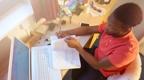 GCSE Biology student using past papers on a laptop and reading a notepad on his desk.