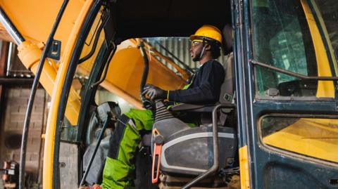 A man in work clothes, including a yellow hard hat, sitting at the controls in the cab of a digger.