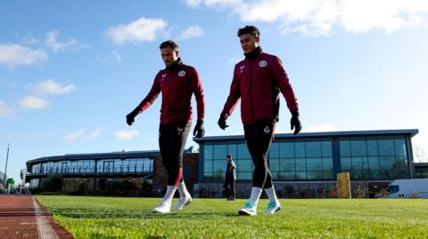 Matty Cash and Ollie Watkins in maroon Aston Villa training kit walking in front of the glass-fronted Bodymoor Heath training ground
