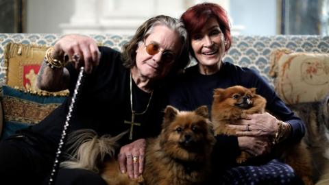 Ozzy Osbourne, who is pictured with long dark grey hair and wearing orange-tinted glasses and chunky gold jewellery, including a cross around his neck, sits on a blue and white patterned sofa with gold and blue cushions next to Sharon Osbourne, who has short bright red hair and is smiling. They are holding two small dogs.