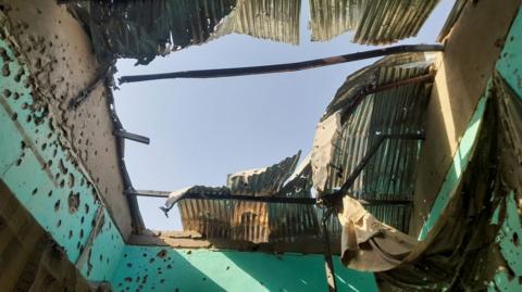 A gaping hole in the roof of a building, with the remains of corrugated iron sheets and bullet marks in the walls.