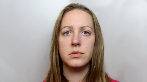 An emotionless Lucy Letby, with long blonde hair and wearing a red jumper, sits in front of a grey wall