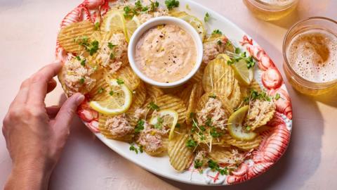 An oval white plate on a pink background filled with crinkle cut crisps and crab meat, with a pot of marie rose sauce and two glasses of lager. 