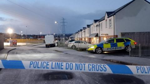 Police tape is in the foreground of the image, behind the tape sits a parked police car, which is in front of a row of houses. A white van and a number of parked car are on the road. the road is icy.