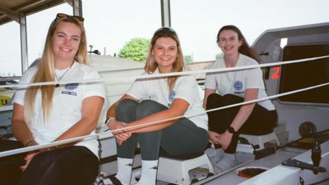 Liv Stocks pictured with Jenna Vincent and Meg Neely. They are in a boat and are wearing white matching t-shirts with a logo. They are all smiling at the camera.