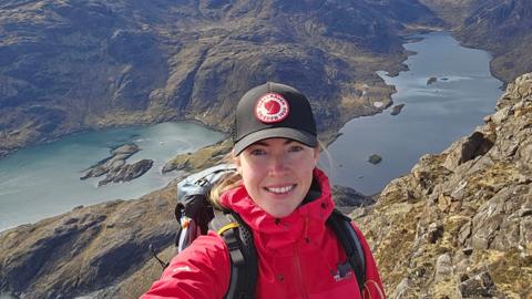 Selfie taken by Louise Percival. She wearing a black cap and red waterproof jacket as she stands on a mountain with two lakes in the background.