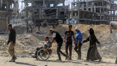 Palestinians walk in front of rubble in Gaza. One of the Palestinians is in a wheelchair, while someone behind him pushes him.