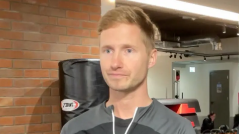 A man in his late twenties wearing a dark athletic top. He is standing in a gym with brick walls, exercise equipment, and a large black punching bag in the background. Additional gym equipment and a seated individual are visible further behind.