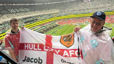 Two football fans in clear rain ponchos hold a large flag in the stands of a football stadium. The white flag features a red St George’s Cross and the Hull City crest. The pitch and seating are visible in the background, with thousands of fans scattered throughout the stadium.