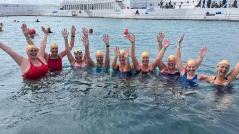 Nine female swimmers wave their arms in the air while paddling around in Penzance's Jubilee Pool. All nine are wearing gold swimming caps.
