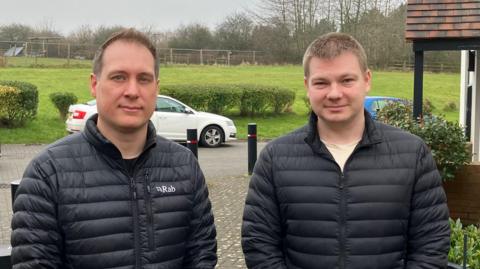 Matthew and Anthony stood in front of the proposed development site. They're both wearing black puffer jackets and smiling slightly. Behind them is a green field with some hedges dividing it from the road. To Anthony's left is the porch of a house, and two cars - one white, one blue - can be see parked on the verge. 