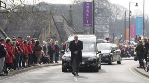 Front view of a funeral cortege. The undertaker is walking in front of a hearse as people - many dressed in red - line the road on either side