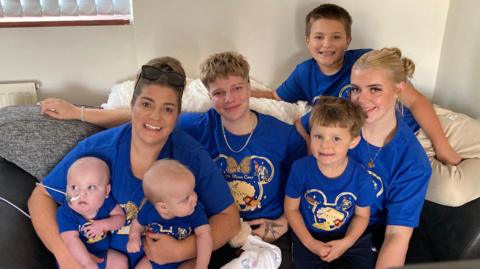 A family pose for a photograph wearng matching blue Disney's The Lion King T-shirts. The mum, who has her dark blonde hair in a bun and has sunglasses on top of her head, is holding two twin baby boys, one of whom has a feeding tube. Also sat on the sofa is two teenage grls - one of whom has very short hair - and two younger boys.