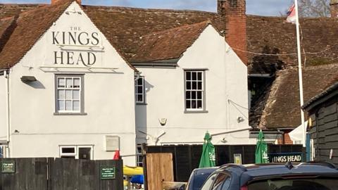 The pub is a white building, with beige roof tiles. The rear of the building has multiple windows on the top half of the building, that have grey ledges. A brown fence is around the building. 