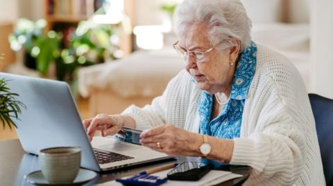 An elderly woman with white hair and glasses is using a laptop while holding a bank card.  There is a mobile phone, a calculator and a cup and saucer on the table.   She is wearing a white cardigan over a blue patterned blouse. 