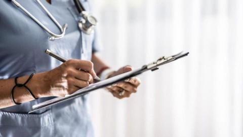 A nurse in blue uniform, holding a clipboard and writing on it. There is a stethoscope around their neck.
