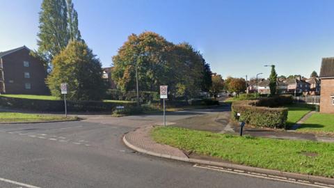 A road in Rotherham showing the entrance to a housing estate and two 20mph signs in view, as well as several trees and gardens 