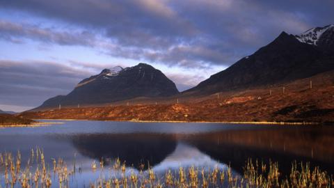 Loch le dà bheinn air a chùlaibh. Tha an adhar sgòthach.