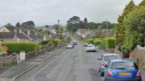 A view down the lane. It is a residential street with cars parked on either side of the roads. 