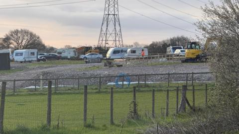 A view of the field. About six caravans are parked on hardstanding along with other vehicles and yellow diggers. There is a pylon in the middle of the field.