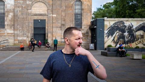 Ross Wilson is standing at the forefront of the picture, with a dark T-shirt on and a gold cross hanging around his neck. He is looking to the right of the picture. Behind him is a converted church, where members of his band are standing or sitting on the steps