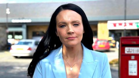 A woman with long black hair, wearing a light blue blazer. She is wearing a silver necklace. The background of the image is blurred, with two cars and a red postbox. 