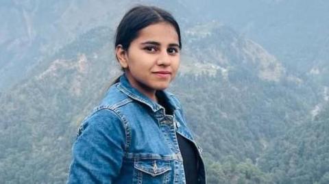 Harshita Brella, who has tied-back black hair and is wearing a denim jacket and a black top. She is posing for a photo in a high spot with tree-covered mountains behind her.