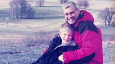 An old picture of Josh and his dad Andrew in the countryside. Both are smiling at the camera, and Josh is leaning back into his dad. They are both wearing warm coats or fleeces. In the background you can see fields and a few sparse trees. 