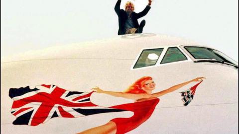 Richard Branson sits on top of a large white plane - only part of the plane can be seen. The windows for the pilots cockpit and underneath where there is the painting of the Scarlet Lady. She is smiling, looking like she is flying forward, wearing a read leotard and holding a billowing union jack flag behind her.