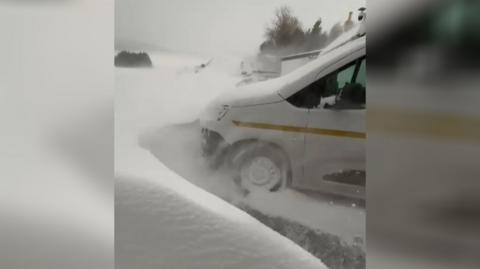 The front of a parked van is surrounded by a high wall of snow, which is still falling.
