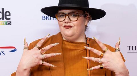 Jayde Adams in an orange blouse and jacket, wearing a black fedora hat. She is holding up her hands, which have long gold and silver nails and a chunky ring on each finger. She is wearing tortoiseshell glasses
